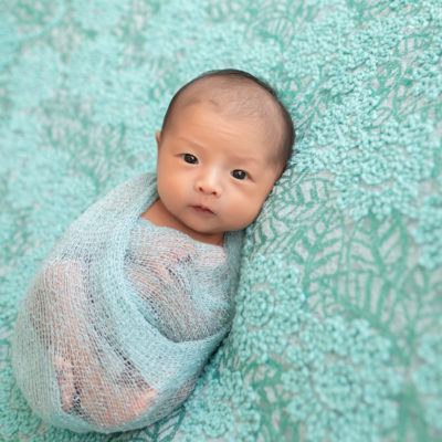 Gainesville newborn portraits baby girl looking straight at camera with her eyes open wrapped in aqua fabric and lying on pale aqua floral blanket