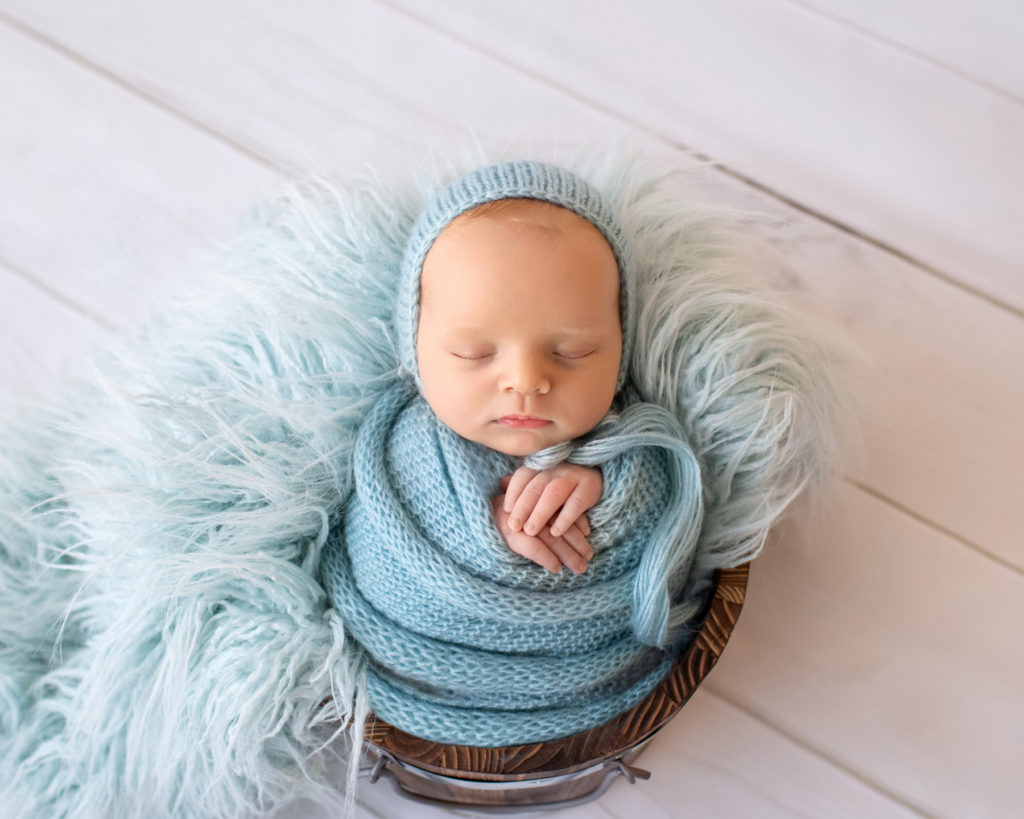 Newborn baby boy Ezra in blue wrap and blue bonnet sound asleep in blue fur stuffed brown bucket Gainesville Florida newborn photographer