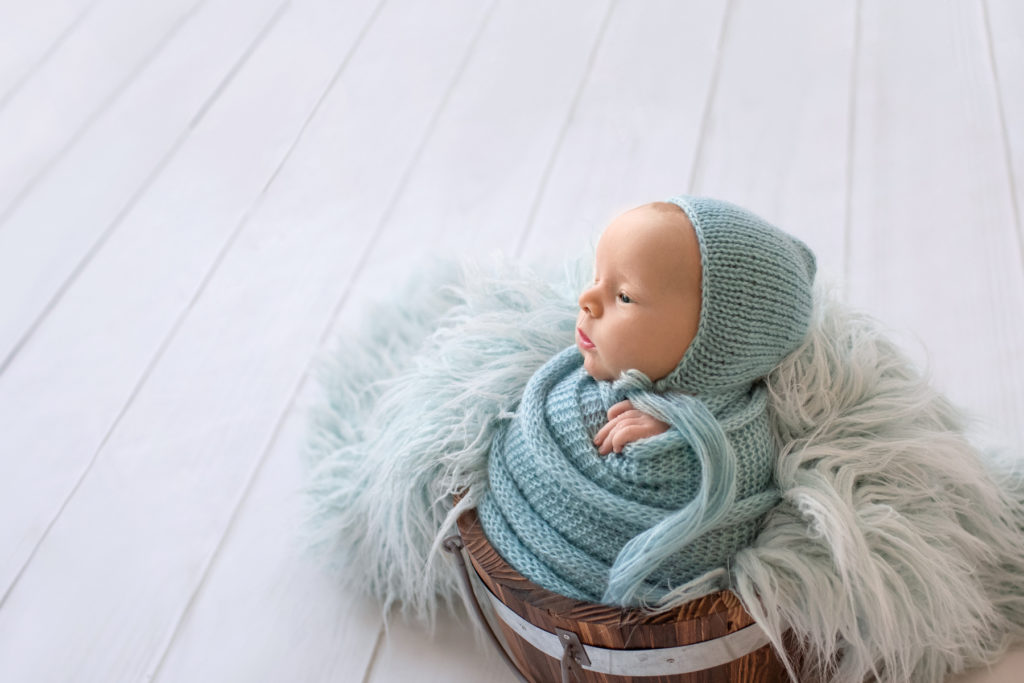 Newborn baby boy Ezra in blue wrap and blue bonnet with eyes wide open in blue fur stuffed brown bucket profile shot Gainesville Florida newborn photographer