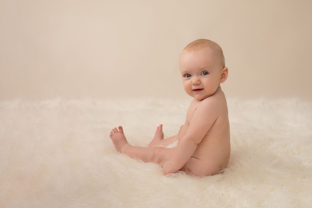 six month naked baby Rachel with eyes locked to camera chin to shoulder like a little baby model posed beautifully on white fur sitting up smiling