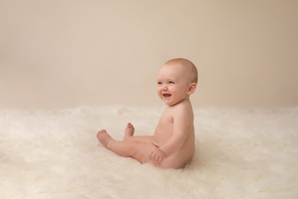 six month naked baby Rachel laughing posed beautifully on white fur sitting up looking at mom