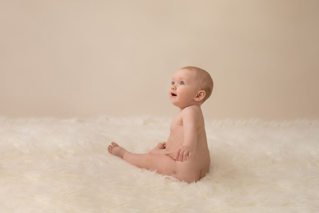 six month naked baby Rachel posed beautifully on white fur sitting up looking up at mom