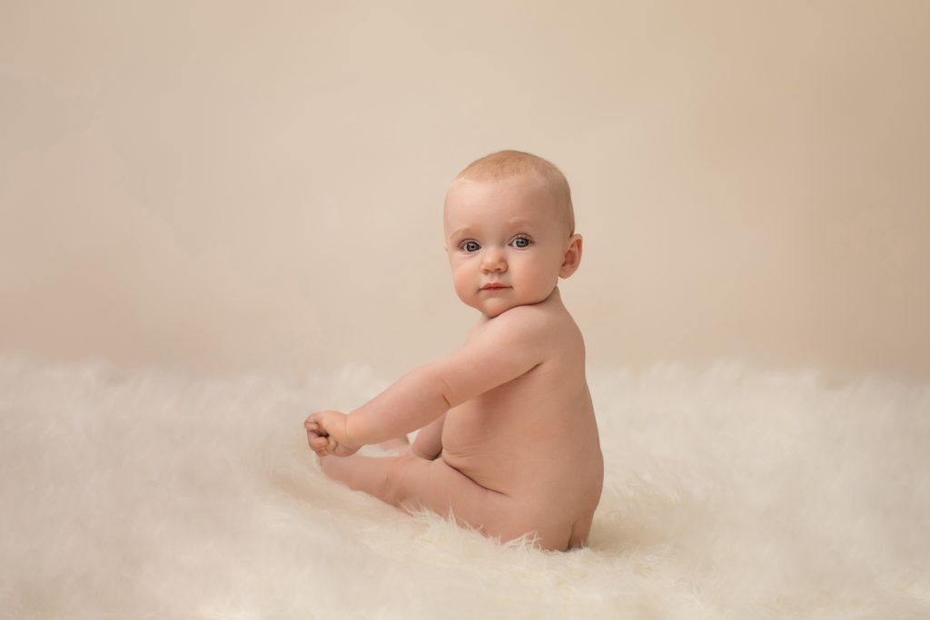 six month naked baby Rachel with eyes locked to camera chin to shoulder like a little baby model posed beautifully on white fur sitting up