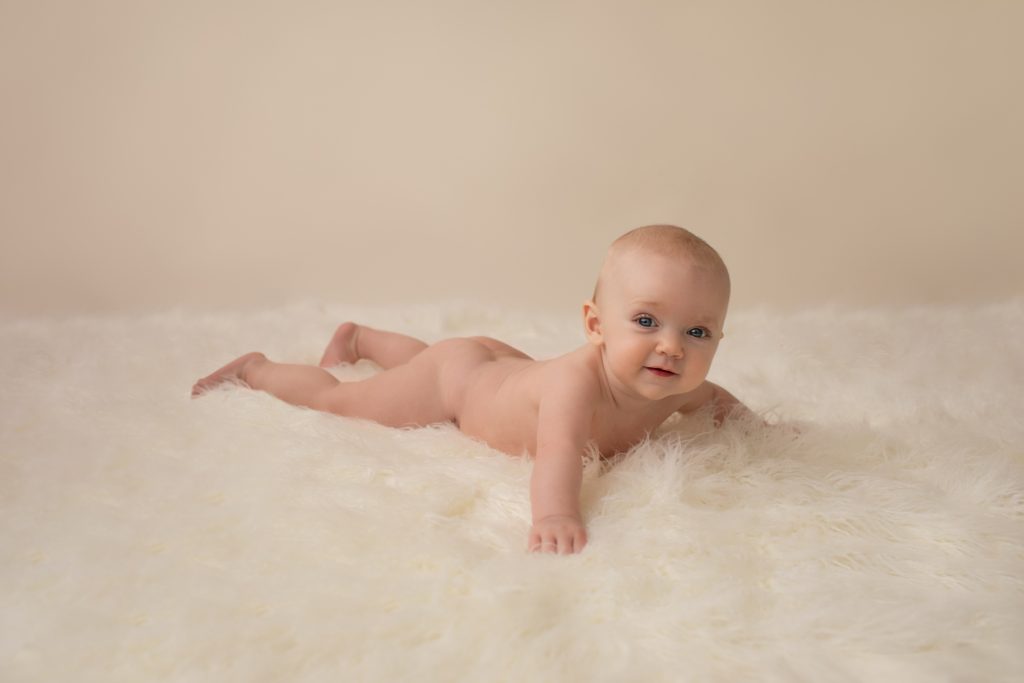 six month naked baby Rachel with soft baby skin grinning at camera on white fur lying on her tummy pushing up on elbows eyes to camera