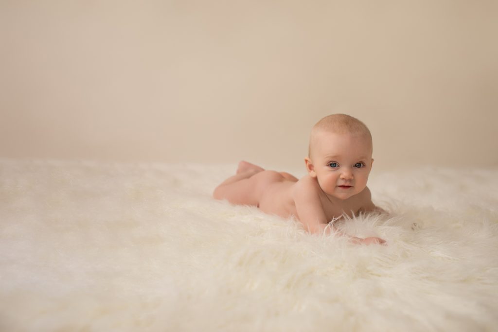 six month naked baby Rachel with soft baby skin posed beautifully on white fur lying on her tummy pushing up on elbows eyes to camera