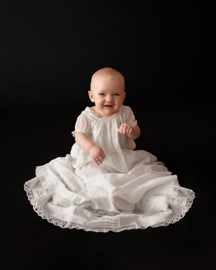 6 month old baby Rachel laughing posed in 205 year old baby christening gown sitting up against a solid black backdrop