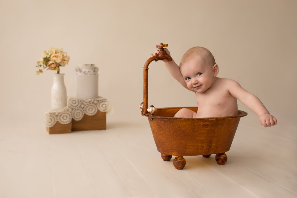 six month naked baby Rachel smiles with personality sitting in brown old fashioned tub playing peek a boo